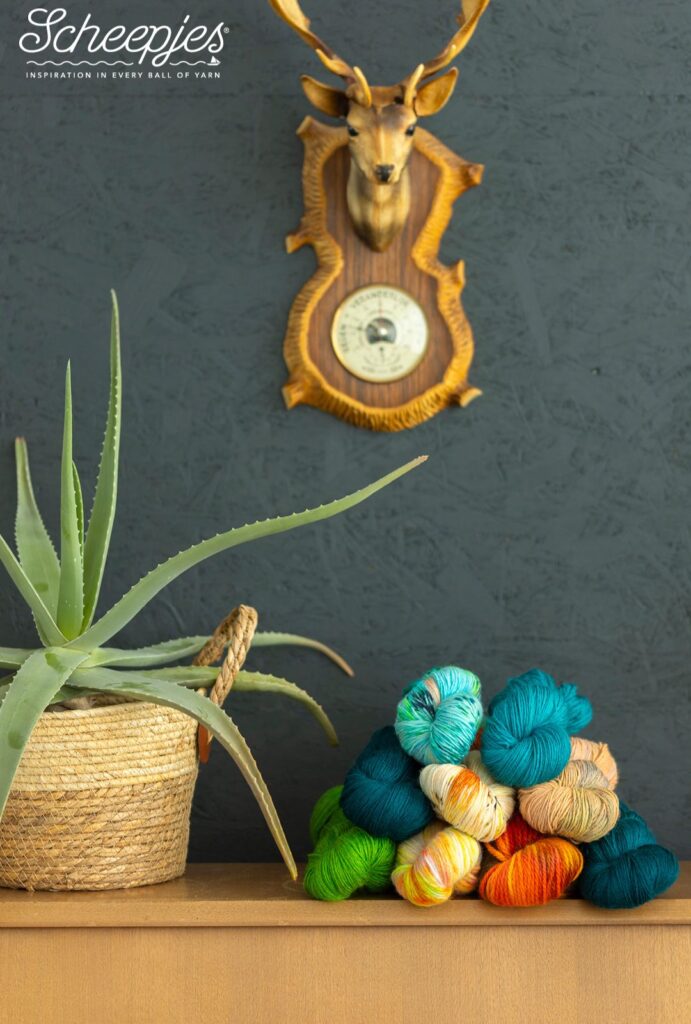 Ontdek de Scheepjes Nakid Series Ongeverfde Garens | Het Wolhuis Een stapel kleurrijke garenstrengen van Scheepjes, liggend op een houten kastje naast een grote aloë vera plant in een gevlochten mand. Op de achtergrond hangt een klassieke wanddecoratie met een hertenhoofd. De strengen zijn in diverse tinten zoals groen, oranje, blauw en beige, en passen perfect bij verschillende creatieve projecten.